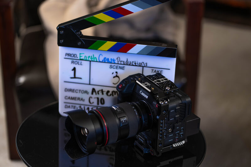 A clapperboard marked 'Earth Coast Productions, Director: Artem' rests behind a Canon C70 cinema camera on a glass-topped side table.
