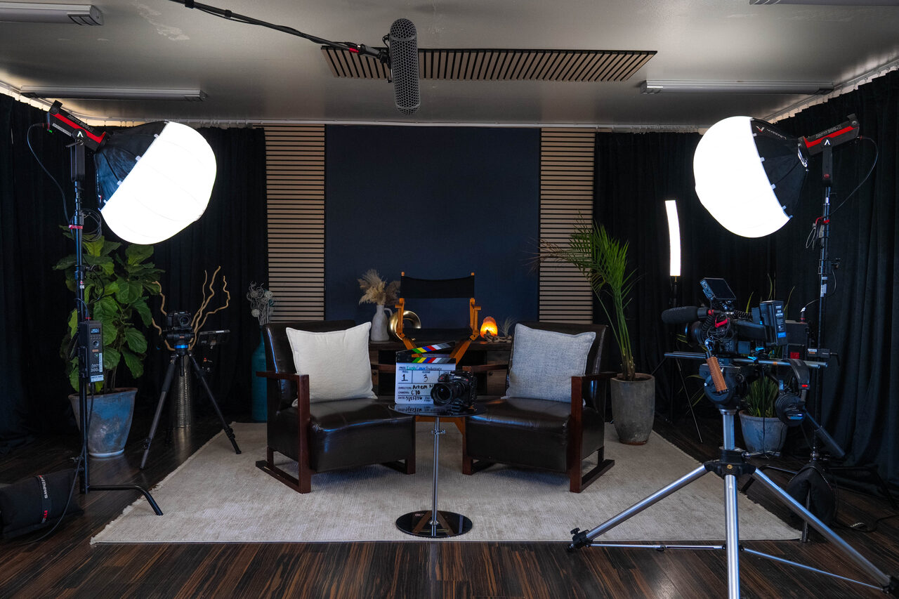 The Earth Coast Studios interview set: two leather armchairs centered between two large softbox lights, a clapperboard and cinema camera on a low stool between them, plants and dark drapes framing the scene.