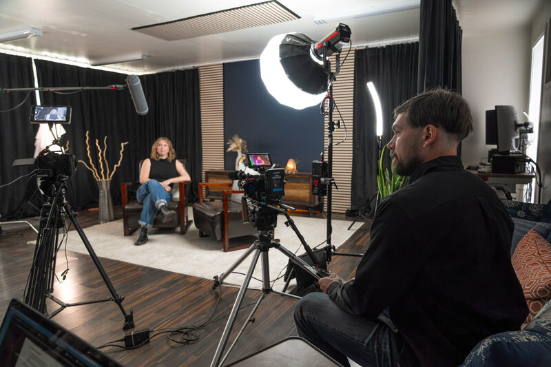 A director sits behind the camera while an interview subject settles into the chairs on the lit studio set.